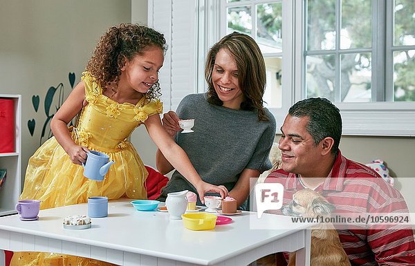 Mädchen im Spielzimmer sitzt am Tisch und serviert Tee vom Spielzeugtee-Set an die Eltern.