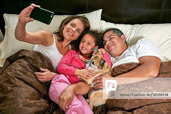 High-Winkel-Ansicht des Mädchens im Bett mit Eltern und Hund mit Smartphone zu nehmen Selfie lächeln