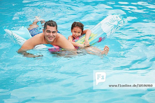 Vater und Tochter im Schwimmbad mit aufblasbarer Matratze und lächelndem Blick auf die Kamera