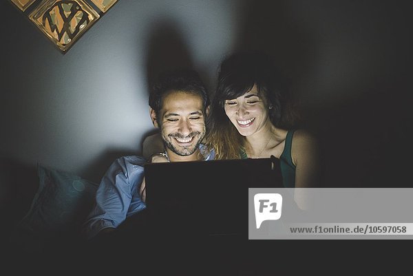 Vorderansicht des jungen Paares  beleuchtet von einem Laptop-Bildschirm  mit lächelndem Blick nach unten.