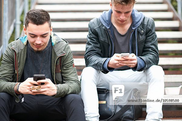 Zwei junge Männer  auf Stufen sitzend  mit Smartphones  im Freien