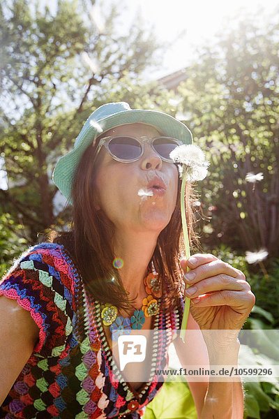 Mature woman wearing sunhat and sunglasses blowing dandelion seeds
