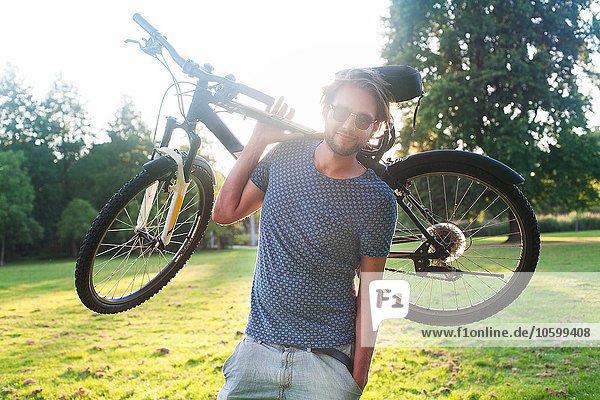 Portrait of young man carrying his bicycle in park at sunset