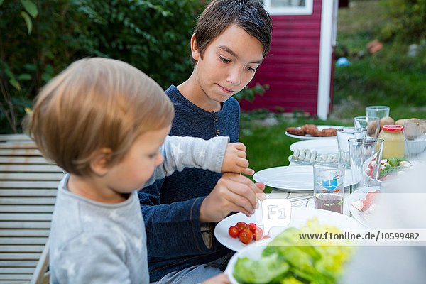 Teenage boy helping female toddler eat food at garden barbecue