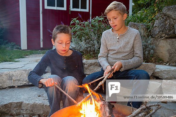 Zwei Jungen mit Stöcken sitzen am Lagerfeuer in der Abenddämmerung.