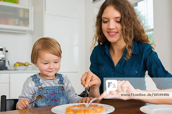 Teenage girl cutting cake for female toddler at kitchen table