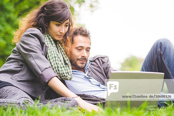 Young couple sitting on grass looking down using laptop computer