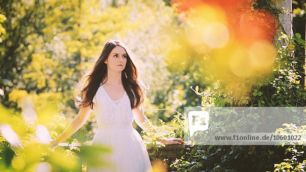 Junge erwachsene Frau in weißem Kleid lehnt an einer Steinmauer im Garten