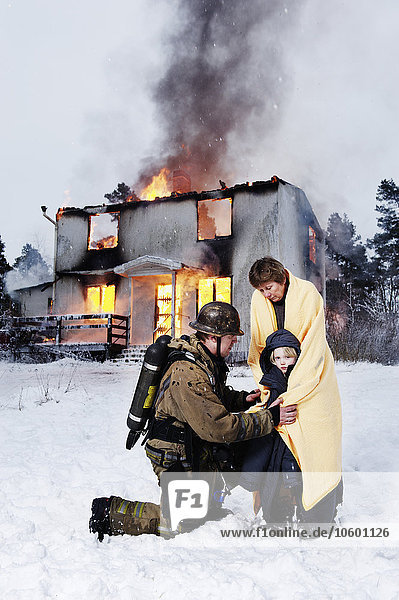 Feuerwehrmann mit geretteter Frau und Mädchen vor einem brennenden Haus