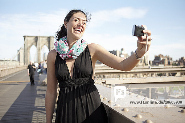 Frau im schwarzen Kleid macht Selfie mit Brooklyn Bridge im Hintergrund