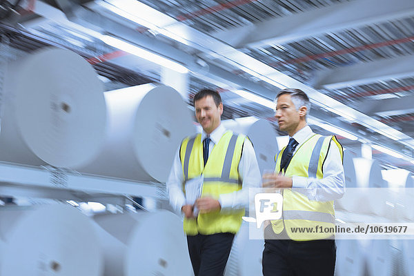 Geschäftsleute in reflektierender Kleidung, die in der Druckerei an großen Papierrollen entlanggehen