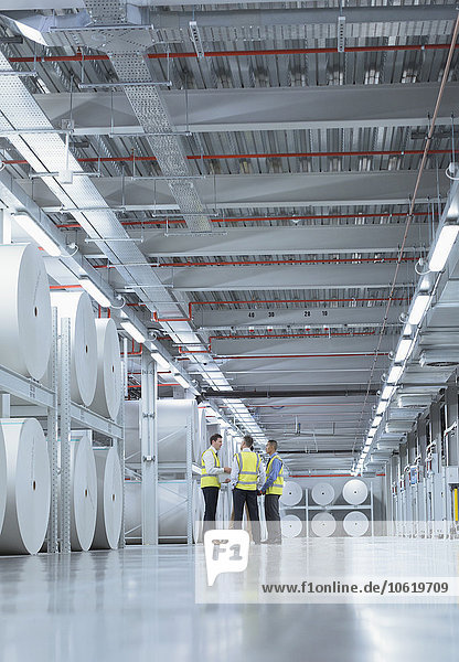 Arbeiter in reflektierender Kleidung reden in der Nähe von großen Papierrollen in der Druckerei