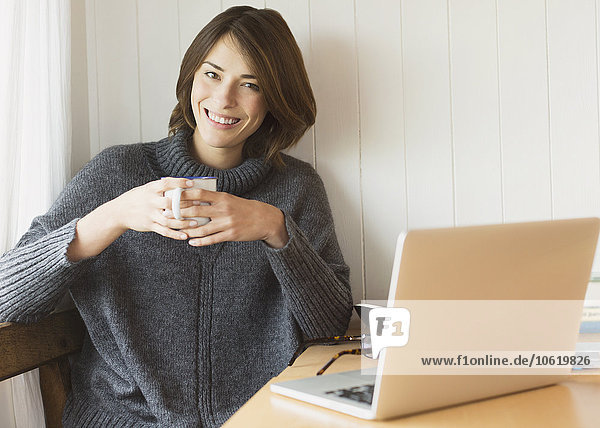 Portrait lächelnde brünette Frau im Pullover beim Kaffeetrinken am Laptop