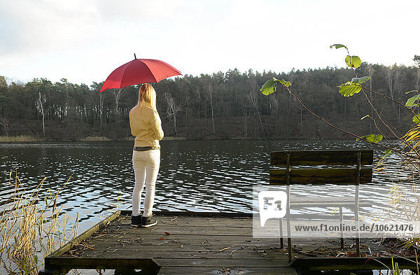 Rückansicht einer blonden Frau auf einem Holzsteg mit rotem Regenschirm und Blick auf einen See.