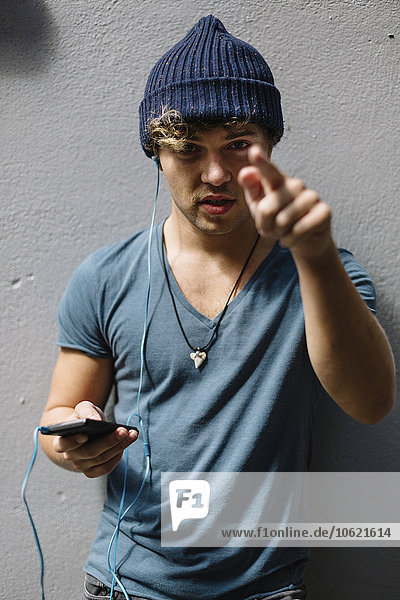 Young man with smartphone and earphones leaning against facade pointing on viewer