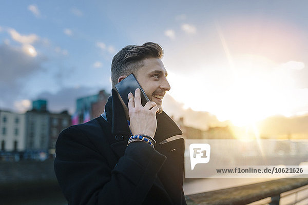 Irland  Dublin  lächelnder junger Geschäftsmann telefoniert mit Smartphone im Gegenlicht