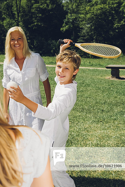 Junge  der mit seiner Schwester Tennis spielt  während seine Mutter im Hintergrund steht.