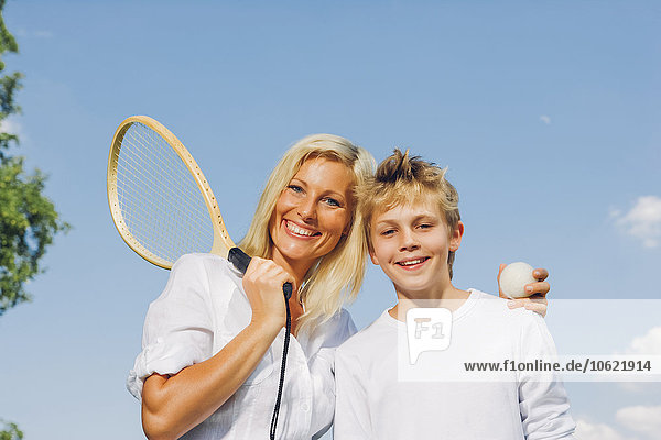 Portrait von glücklicher Mutter und Sohn mit Tennisschläger und Ball am blauen Himmel
