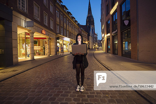 Deutschland  Münster  Portrait einer jungen Frau mit Laptop in der Stadt am Abend