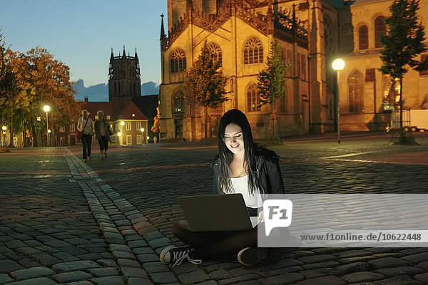 Deutschland  Münster  junge Frau mit Laptop am Abend in der Stadt auf dem Bürgersteig sitzend
