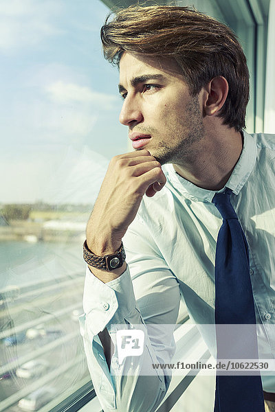 Porträt eines jungen Geschäftsmannes mit Hand am Kinn durchs Fenster schauend