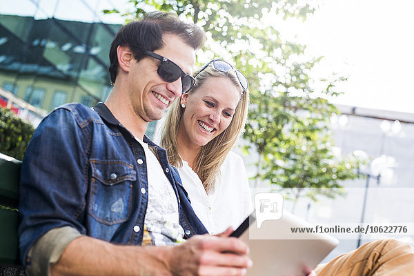 Happy young couple sitting on bench looking at digital tablet