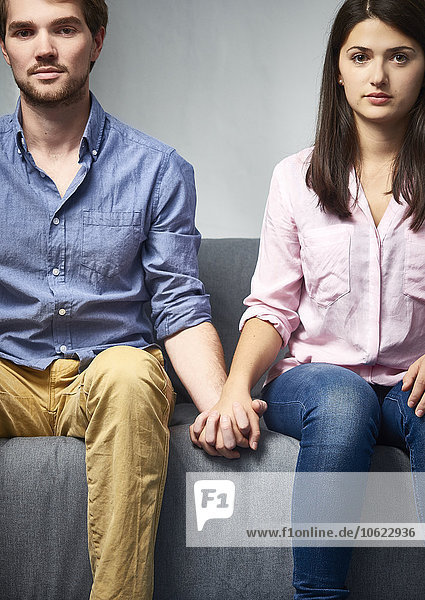 Serious young couple sitting on couch holding hands