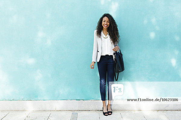Porträt einer lächelnden jungen Geschäftsfrau mit Ledertasche vor einer blauen Wand stehend