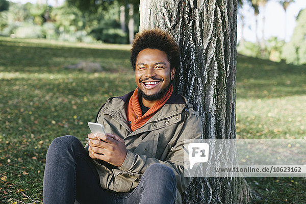 Portrait of smiling young man with smartphone leaning against tree trunk