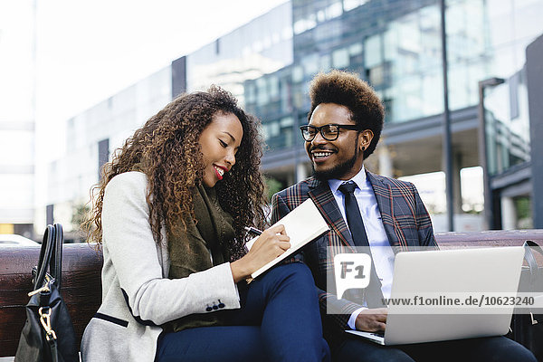 Two smiling young business people with laptop and notebook on bench