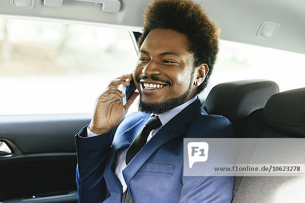 Lächelnder Geschäftsmann sitzt auf dem Rücksitz eines Autos und telefoniert mit dem Smartphone.