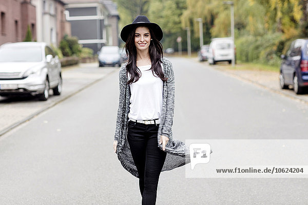 Porträt einer lächelnden jungen Frau mit schwarzem Hut  die auf einer Straße läuft.