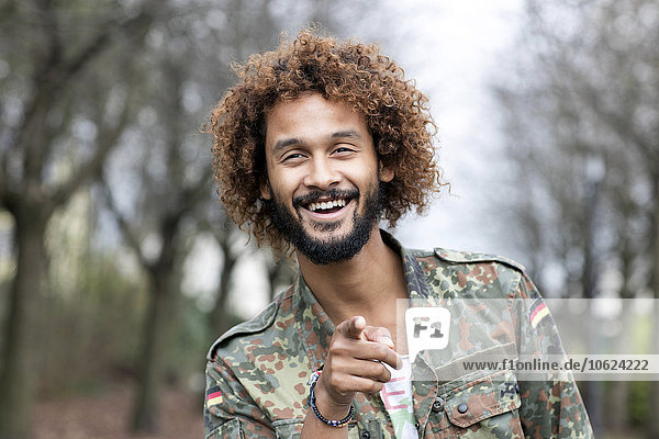 Porträt eines lächelnden Mannes mit gefärbten Ringeln im Camouflage-Shirt  das auf den Betrachter zeigt.