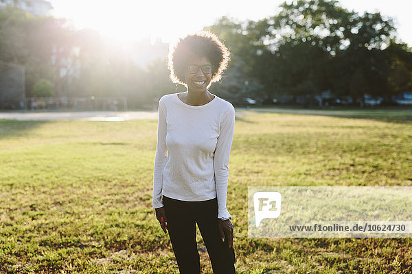 Lächelnde junge Frau steht auf einer Parkwiese im Gegenlicht