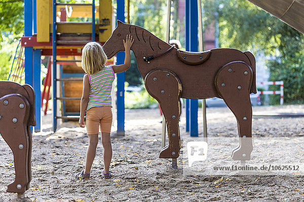 Kleines Mädchen auf dem Spielplatz stehend streichelndes Holzpferd