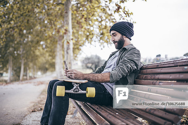 Spanien  Tarragona  junger Mann sitzt auf einer Bank mit seinem Longboard und nimmt einen Selfie mit Smartphone.