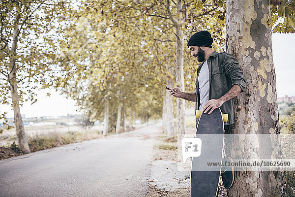 Spain  Tarragona  young man with longboard leaning against tree trunk looking at his smartphone