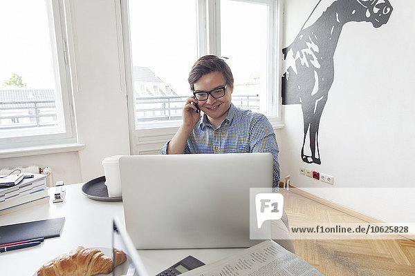 Smiling young businesssman with laptop and cell phone in a modern office