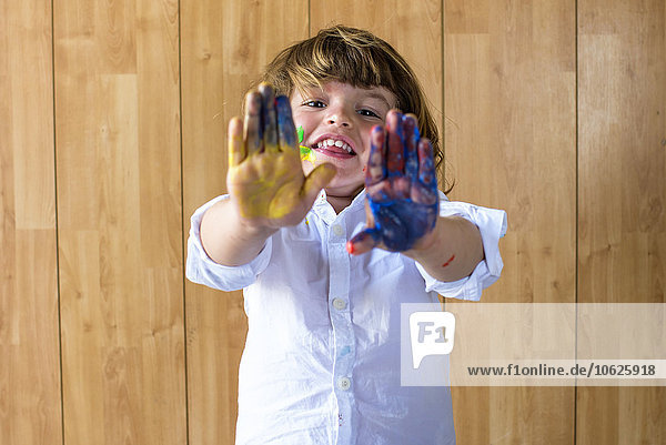 Porträt eines lächelnden kleinen Jungen  der seine Handflächen voller Fingerfarben zeigt.