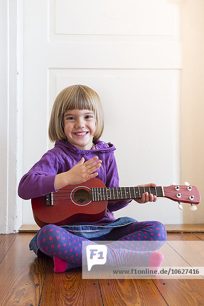 Portrait of little girl sitting on wooden floor playing ukulele