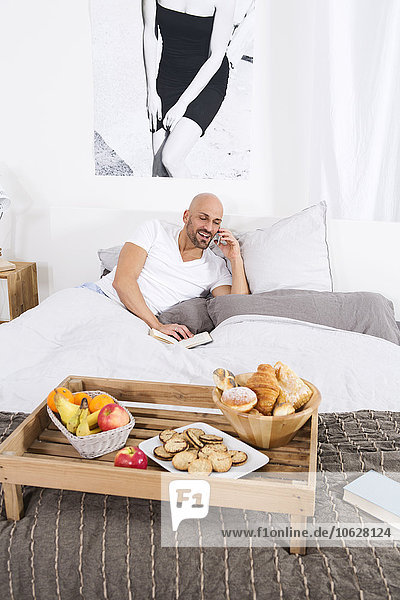 Erwachsener Mann mit Büchern und Frühstück im Bett  Telefongespräch