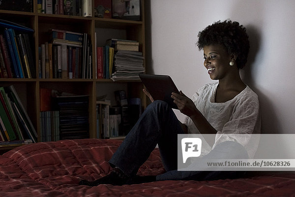Junge Frau sitzt zu Hause auf ihrem Bett und benutzt ein digitales Tablett.