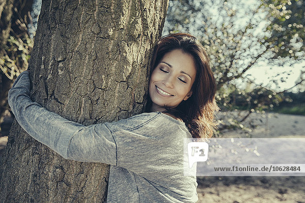 Porträt einer lächelnden Frau mit geschlossenen Augen  die einen Baum umarmt.