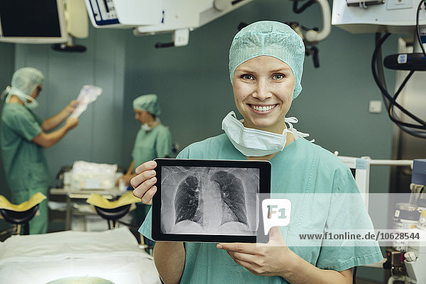 Lächelnde Chirurgin im Operationssaal mit einem digitalen Tablett mit Thorax-Röntgenbild