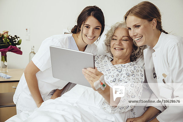 Seniorin im Krankenhaus zeigt Ärztin und Krankenschwester etwas auf digitalem Tablett