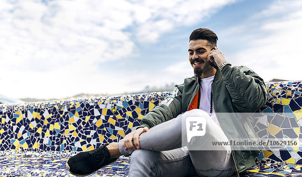Portrait of a stylish young man sitting on tiled bench telephoning with smartphone