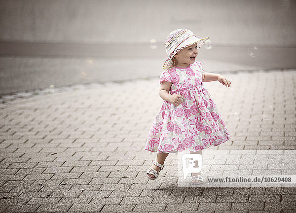 Fröhliches blondes kleines Mädchen mit Hut und Sommerkleid mit Blumenmuster  das auf dem Bürgersteig tanzt.