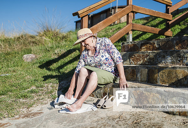 Seniorin sitzt auf einer Treppe und wischt sich den Sand von den Füßen.
