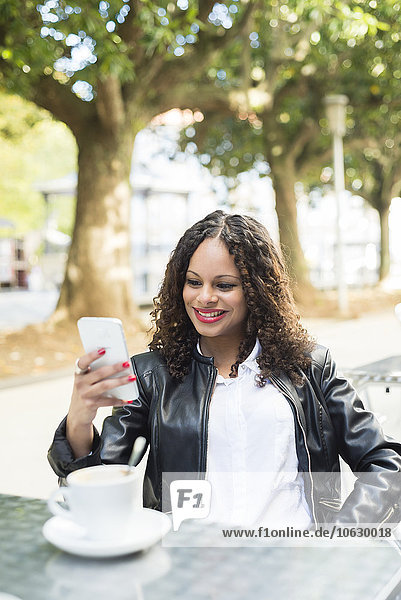 Porträt einer lächelnden jungen Frau  die in einem Straßencafé sitzt und ihr Smartphone ansieht.