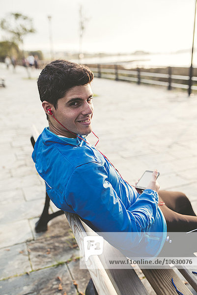 Lächelnder Athlet sitzt nach dem Training auf der Bank und hört Musik vom Smartphone.